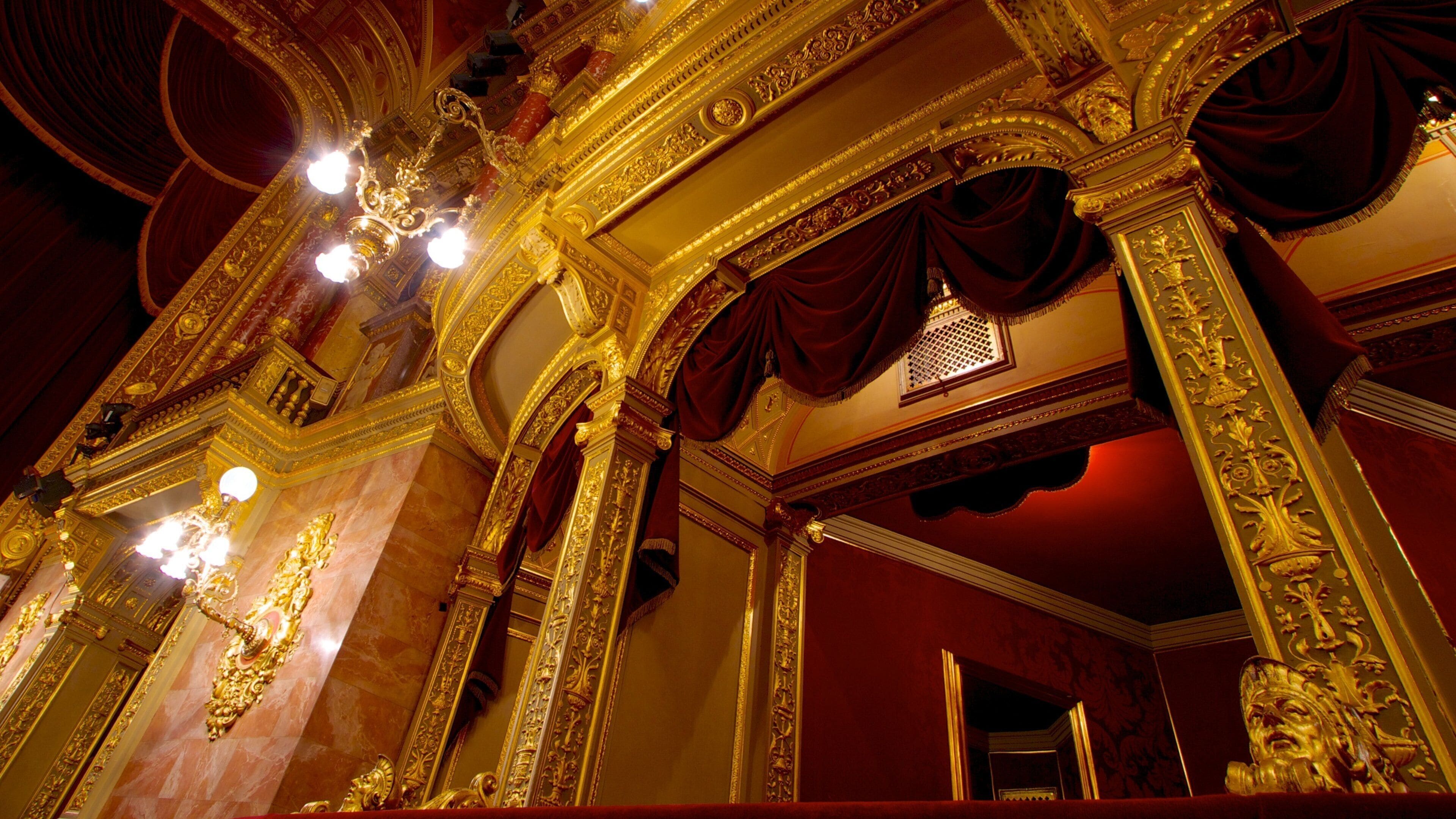 An ornate theater interior with gold detailing, red curtains, and a chandelier.