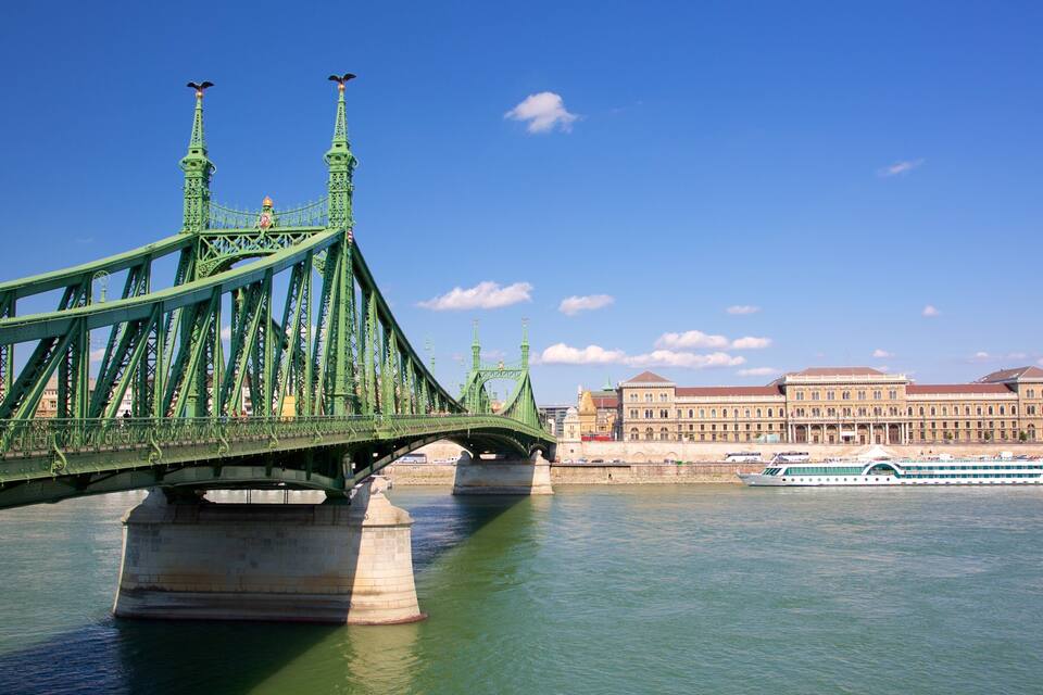Un pont verd amb detalls ornamentals, un edifici gran amb teulada vermella i un riu.