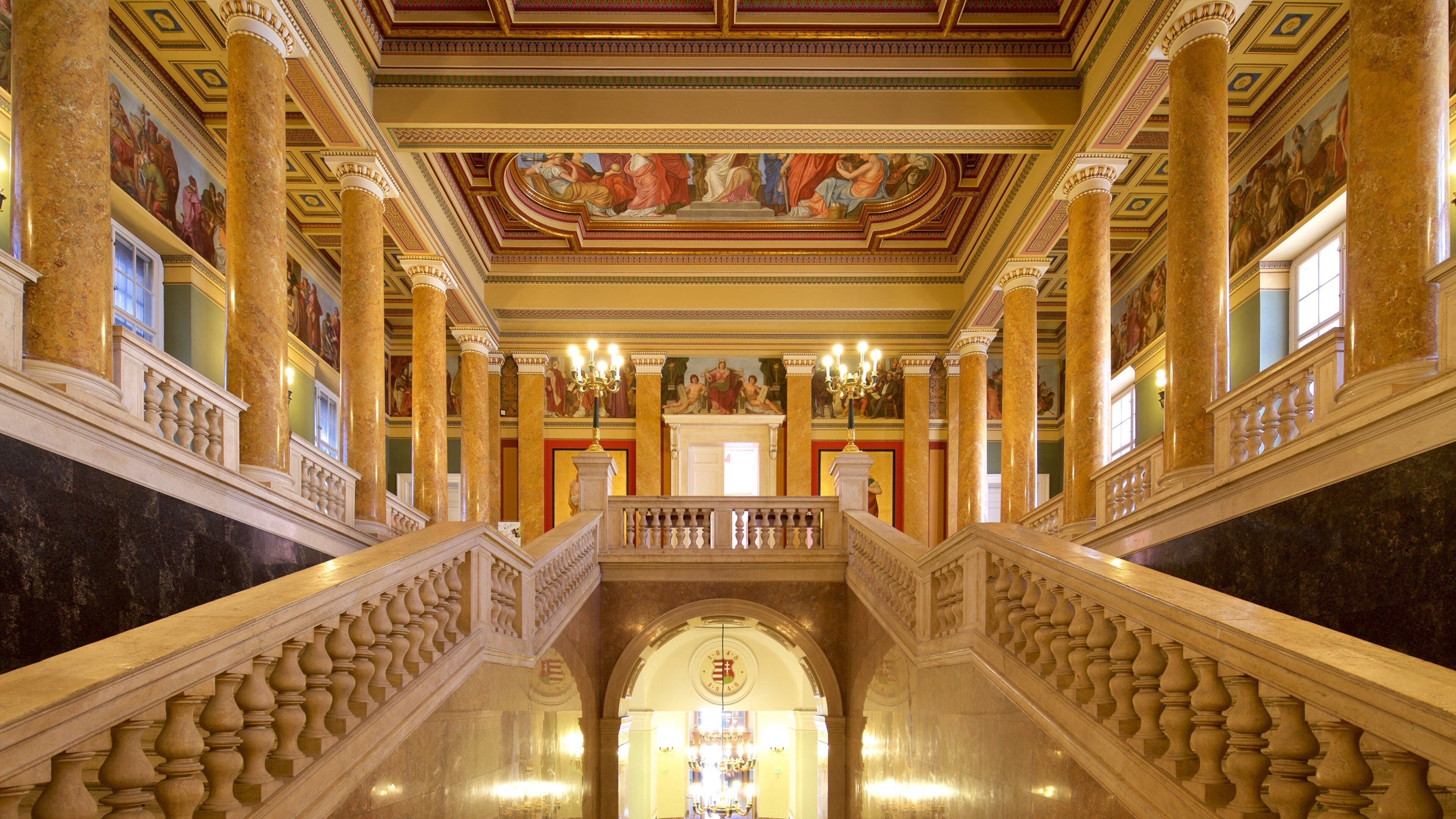 A grand staircase with ornate balustrades, large columns, and a ceiling fresco.