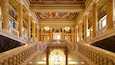 Un magnifique escalier orné de balustrades travaillées, de grandes colonnes et d’une fresque au plafond.