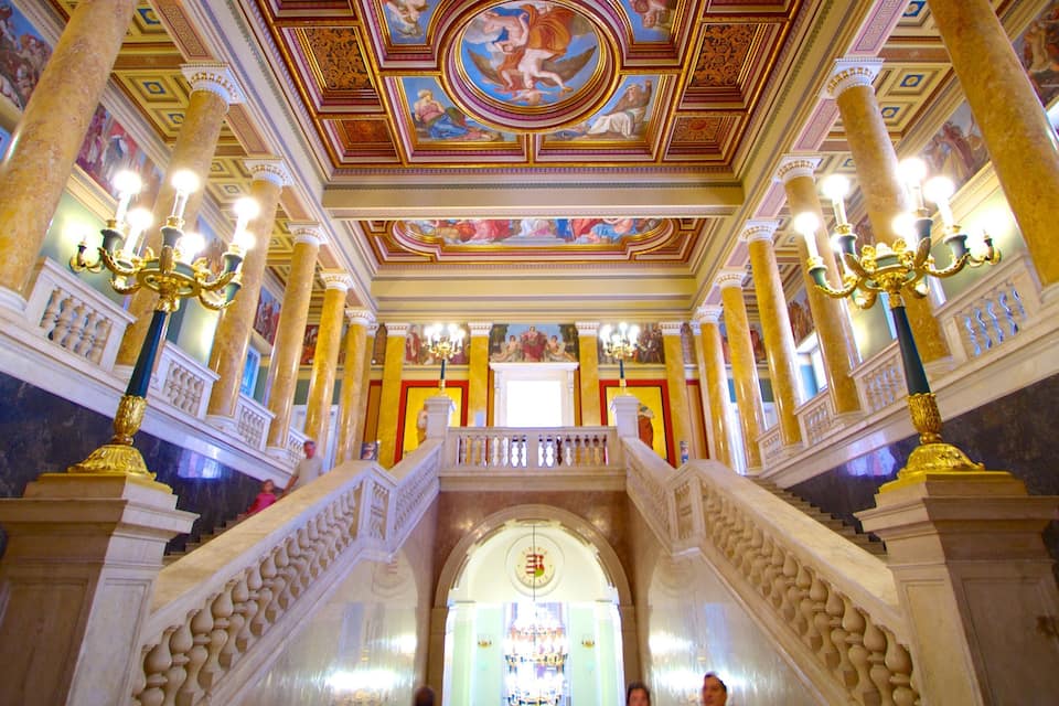 A grand staircase with ornate railings and columns, leading to a brightly lit area with a large chandelier and a ceiling fresco.