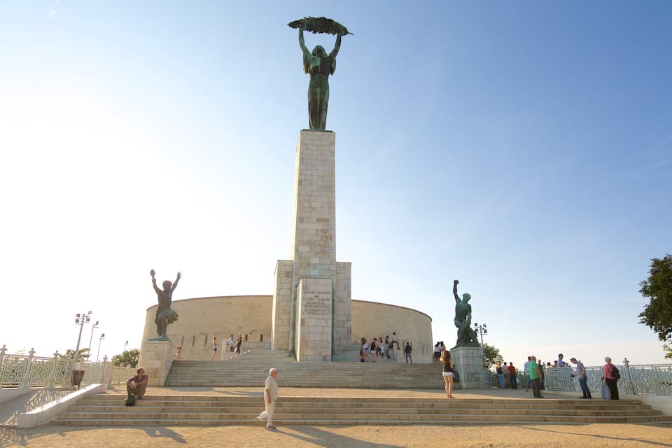 Ein Denkmal mit einer zentralen Statue einer Person, die einen Regenschirm hält, umgeben von kleineren Statuen und einem kreisförmigen Gebäude mit Inschriften.