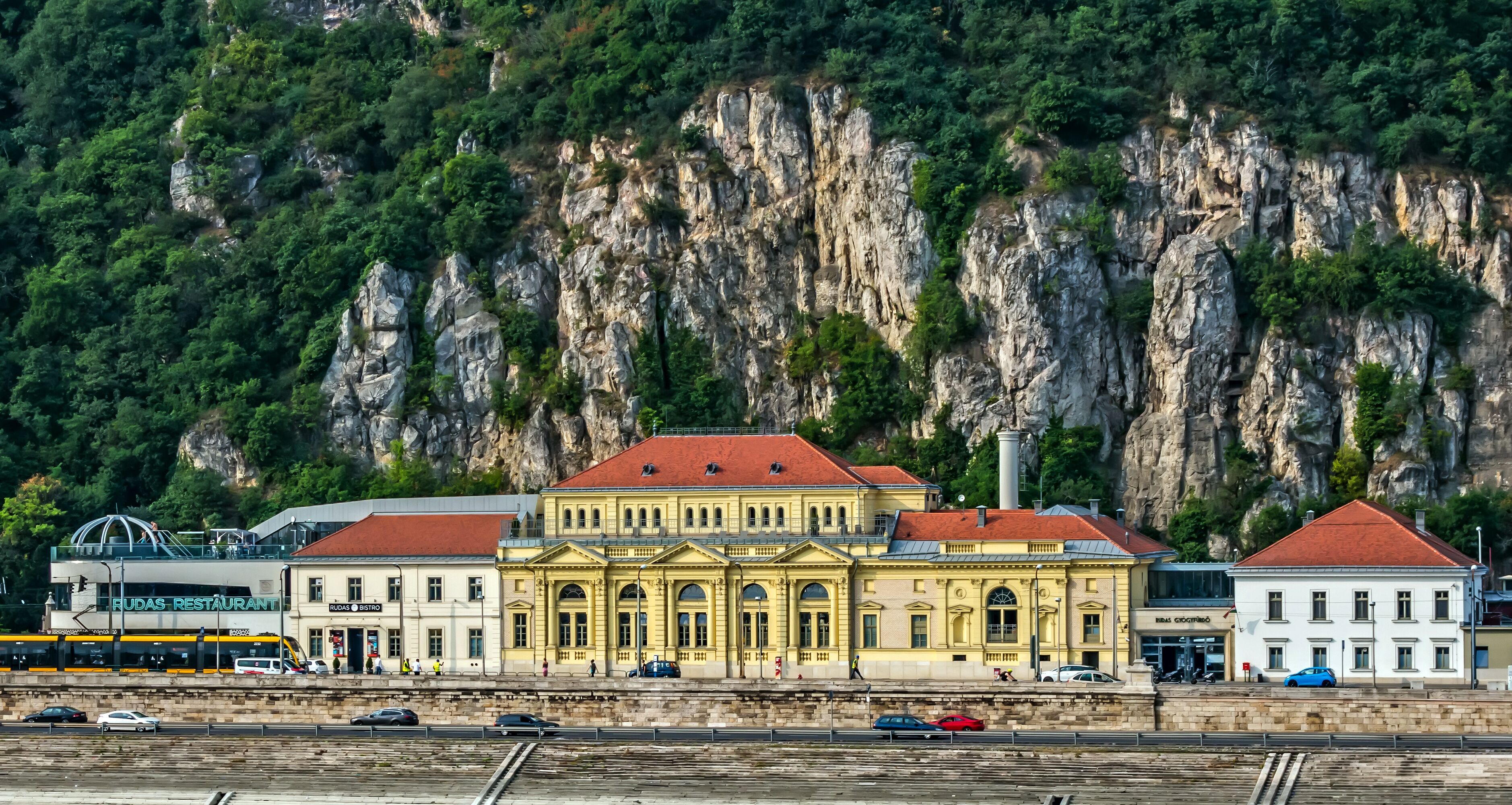 A historic building with a yellow facade and red roof, situated by a railway track with a mountainous backdrop.