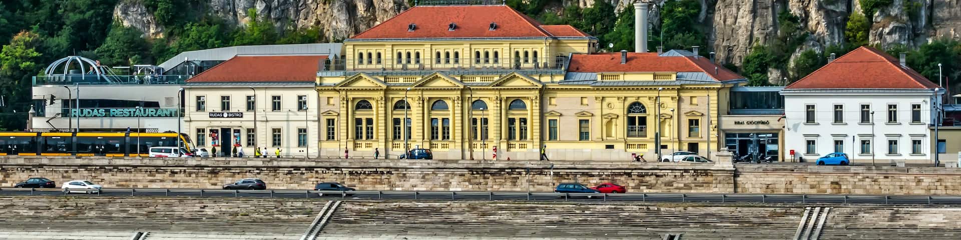 A historic building with a yellow facade and red roof, situated by a railway track with a mountainous backdrop.