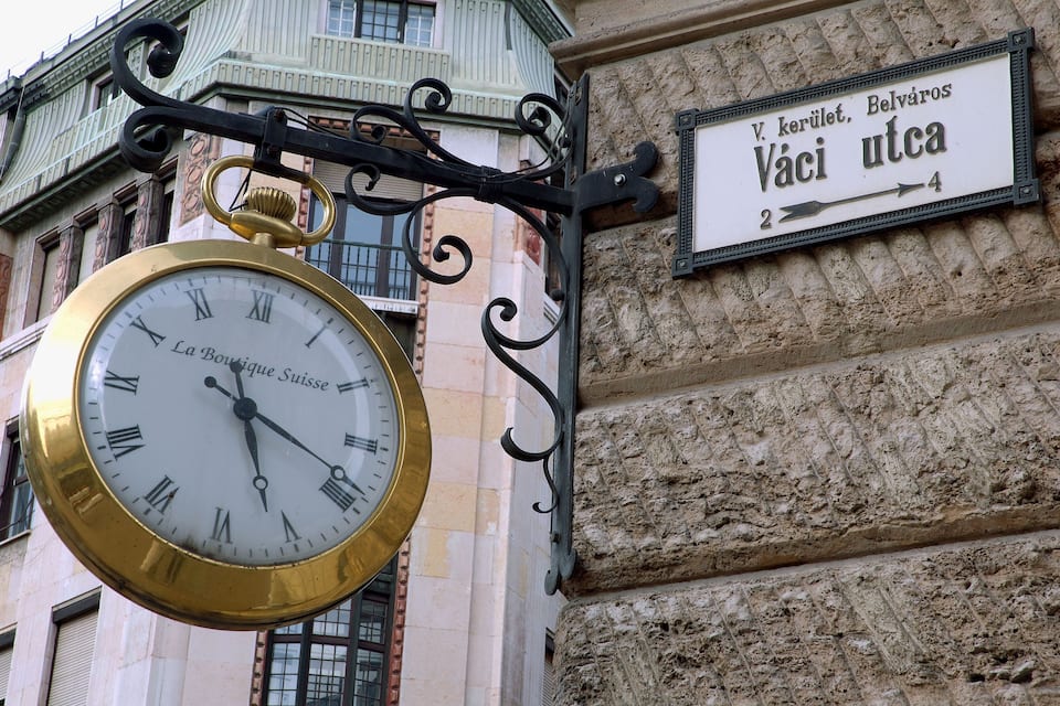 A large gold clock with Roman numerals, a street sign, and a building facade.