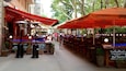 Un café en plein air avec des parasols rouges, des tables et des chaises à l’extérieur, et un sol pavé de briques.