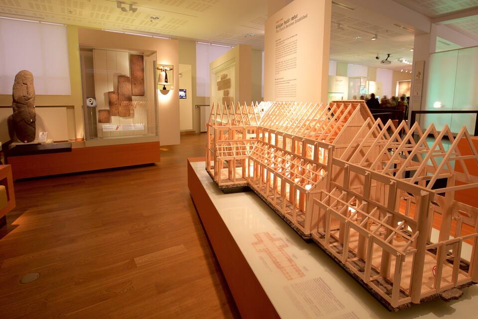 Une exposition de musée comprenant une maquette en bois d’une structure, des vitrines présentant des objets anciens et des panneaux d’information.