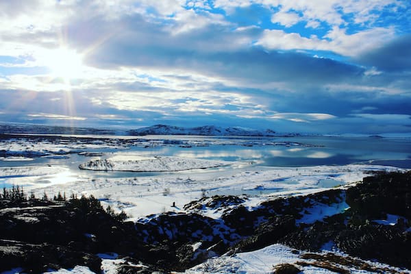 A snowy landscape with a lake, mountains, and a partly cloudy sky.