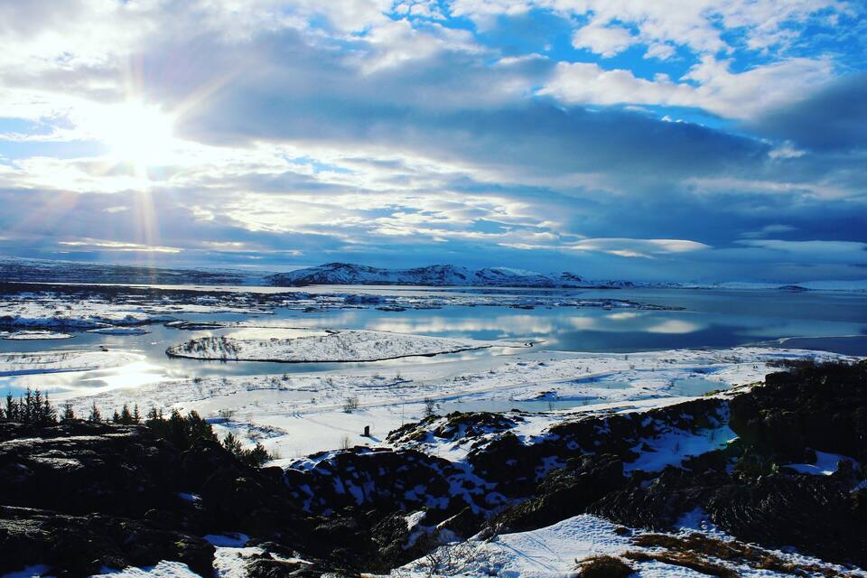 A snowy landscape with a lake, mountains, and a partly cloudy sky.