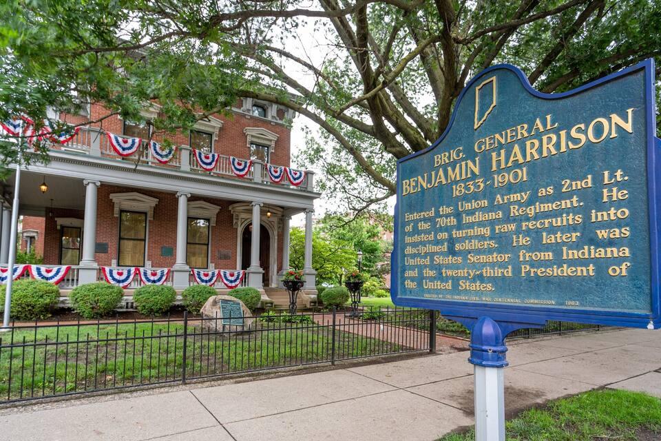 A historical marker for General Benjamin Harrison in front of a brick building with a porch and American flags.