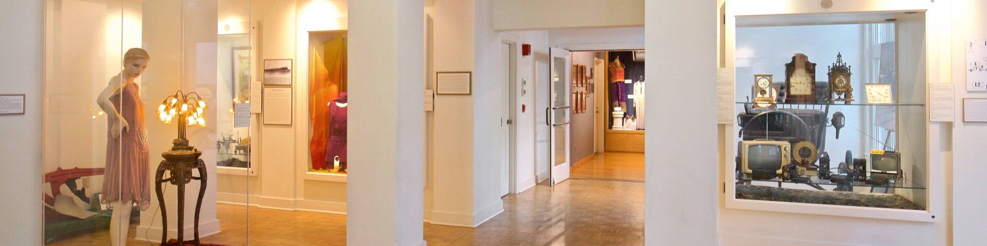 A museum interior with wooden floors, white walls, and various exhibits including mannequins, clocks, and framed pictures.
