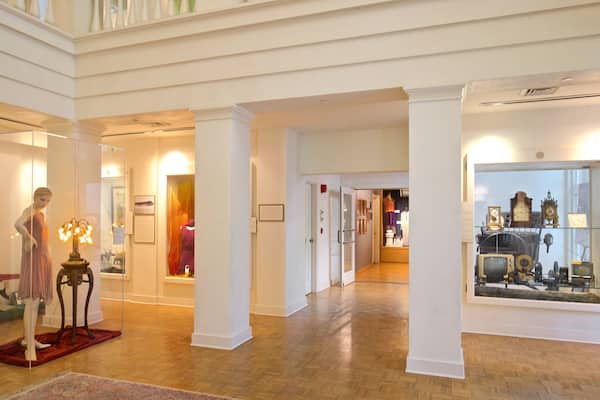 Interior de museu com pisos de madeira, paredes brancas e diversas exposições, incluindo manequins, relógios e quadros emoldurados.