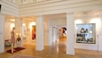 A museum interior with wooden floors, white walls, and various exhibits including mannequins, clocks, and framed pictures.