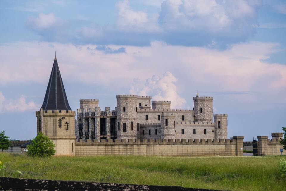 A large castle with multiple towers and a pointed roof, surrounded by a grassy field and a stone wall.