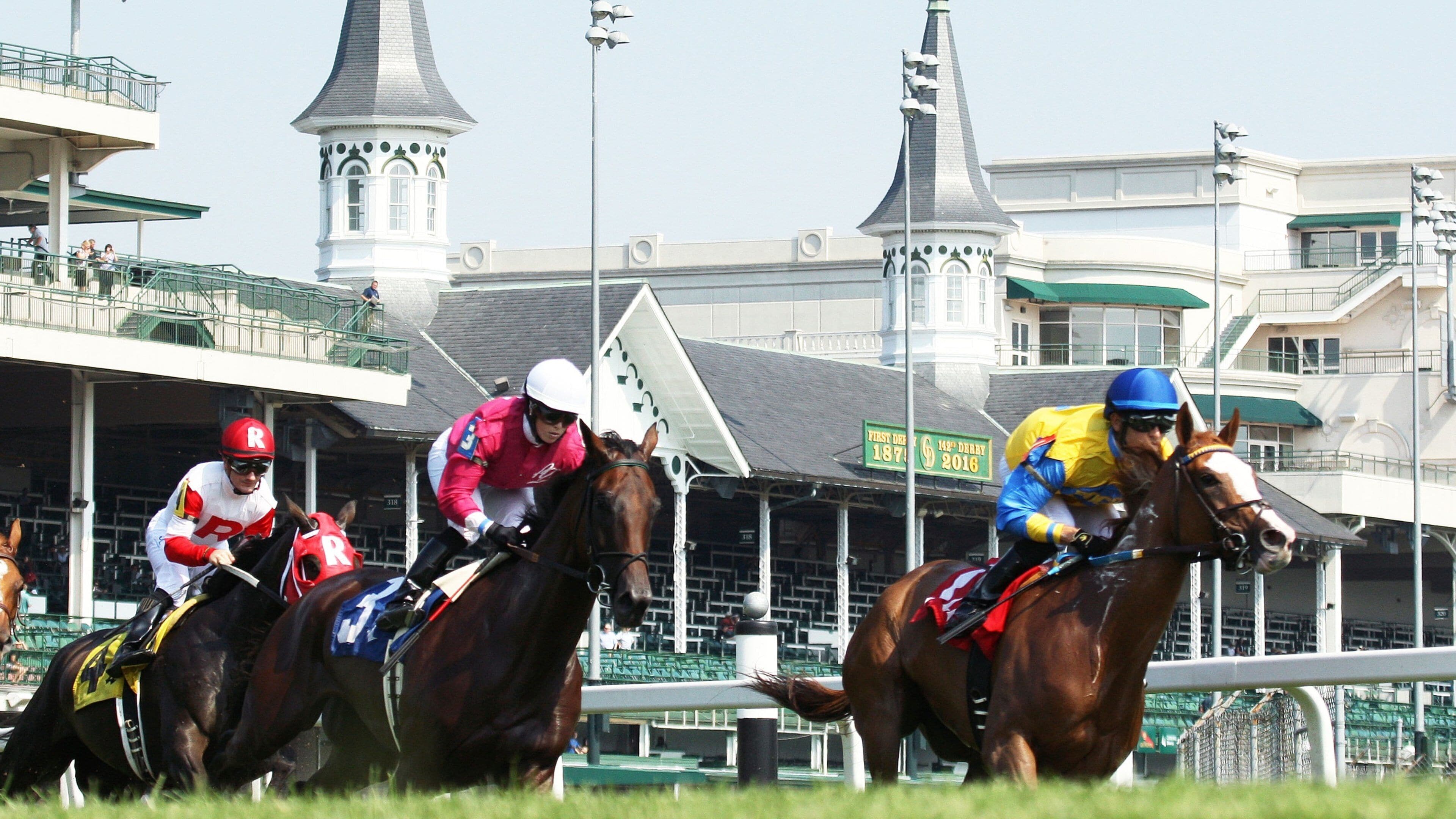 Jockeys on horses racing at a track with distinctive white towers in the background.