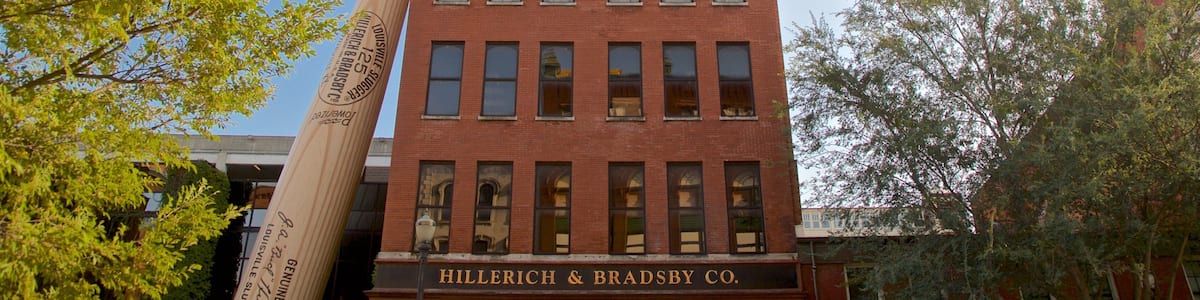 A red brick building with a large wooden baseball bat leaning against it. The building has multiple windows and a sign that reads "Hillerich & Bradsby Co."