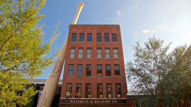 A red brick building with a large wooden baseball bat leaning against it. The building has multiple windows and a sign that reads "Hillerich & Bradsby Co."