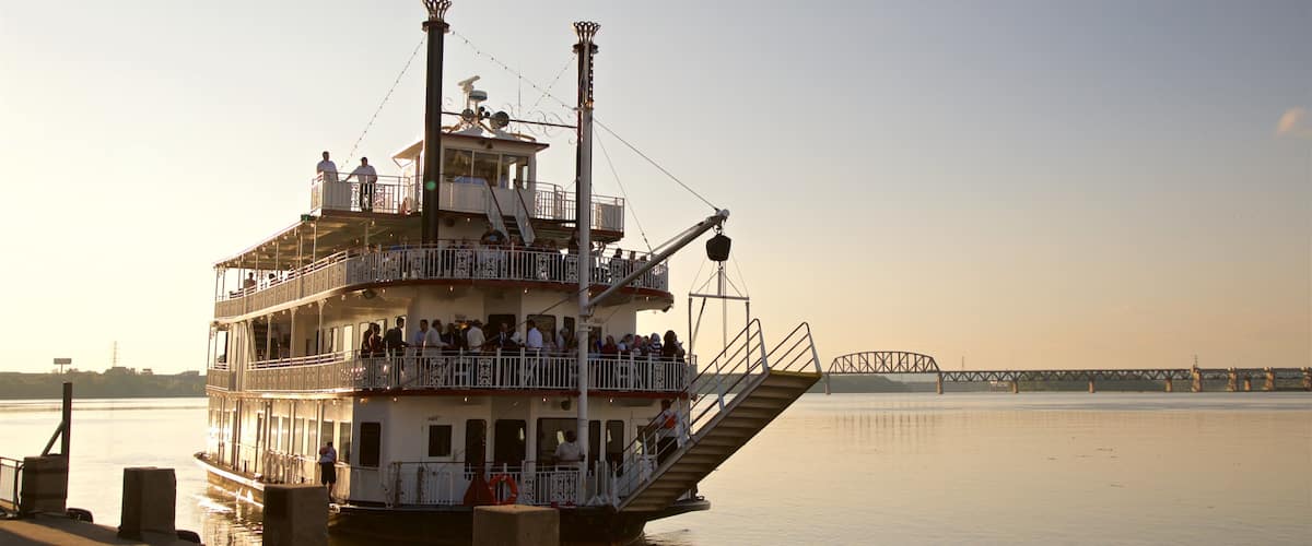 A multi-deck riverboat docked at a pier with a bridge in the background.