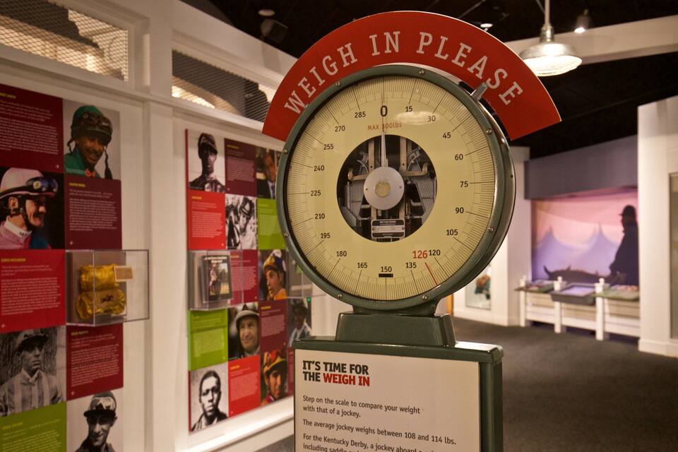 A scale with a sign that reads "WEIGH PLEASE" and a display of jockeys' weights.