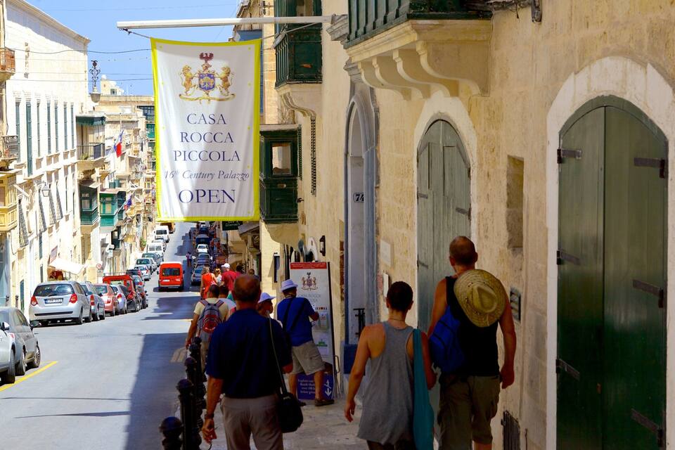 Eine Straße mit parkenden Autos und Fußgängern. Ein Banner hängt an einem Gebäude und zeigt die Aufschrift 'Casa Rocca Piccola 16th Century Palazzo Open'.