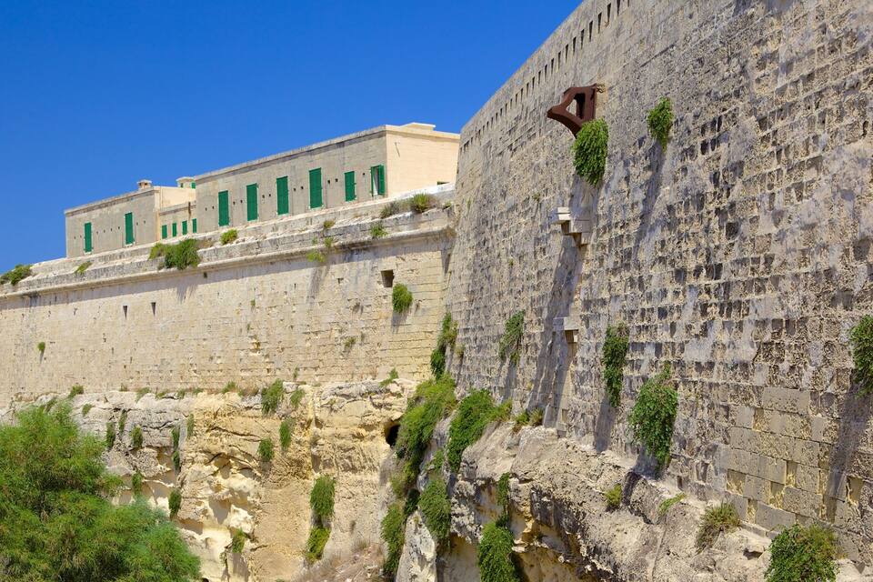 Un muro de piedra histórico con vegetación, un edificio con contraventanas verdes y un cielo azul despejado.