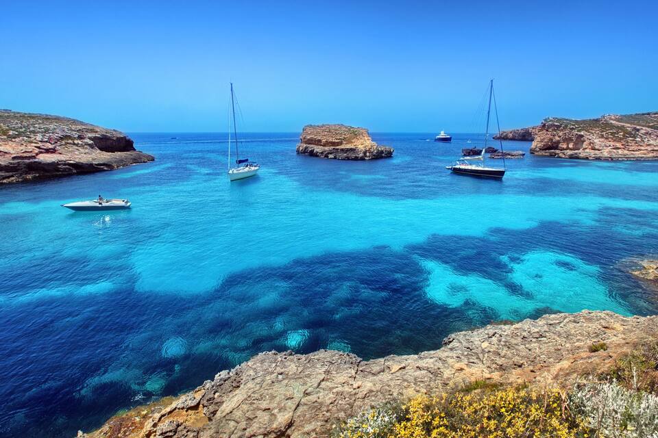 A coastal scene with clear turquoise waters, rocky outcrops, and several sailboats anchored near the shore.