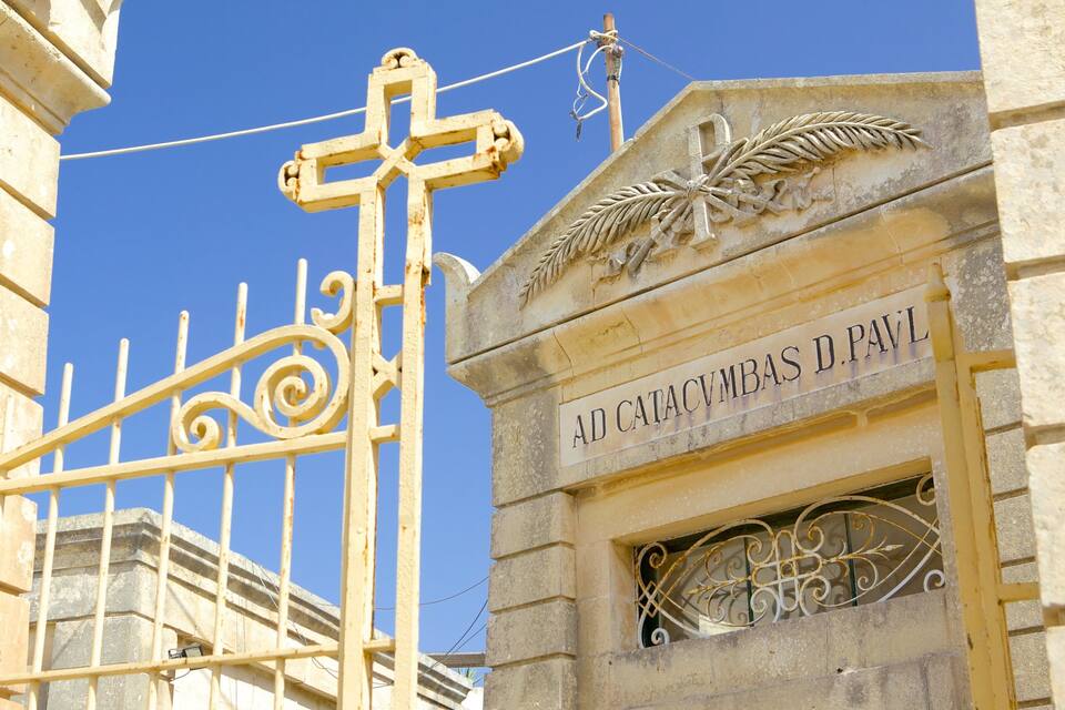 Una reixa de ferro forjat amb una creu, un edifici de pedra amb una fulla gravada i text, i un cel blau clar.