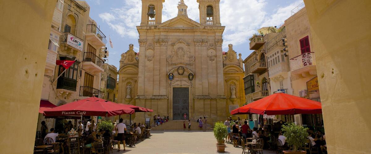 A historic church with two bell towers and a central dome, surrounded by cafes and outdoor seating.