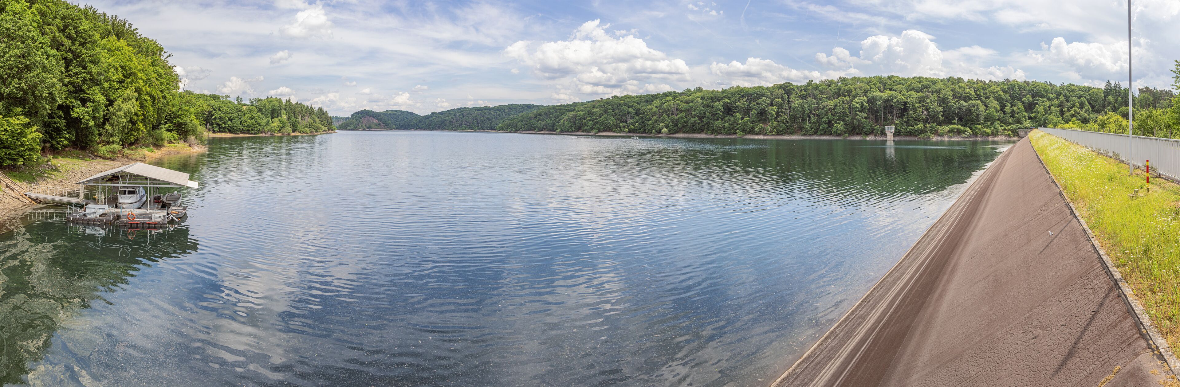 A calm lake with a dock, a dam, and a forested hillside.