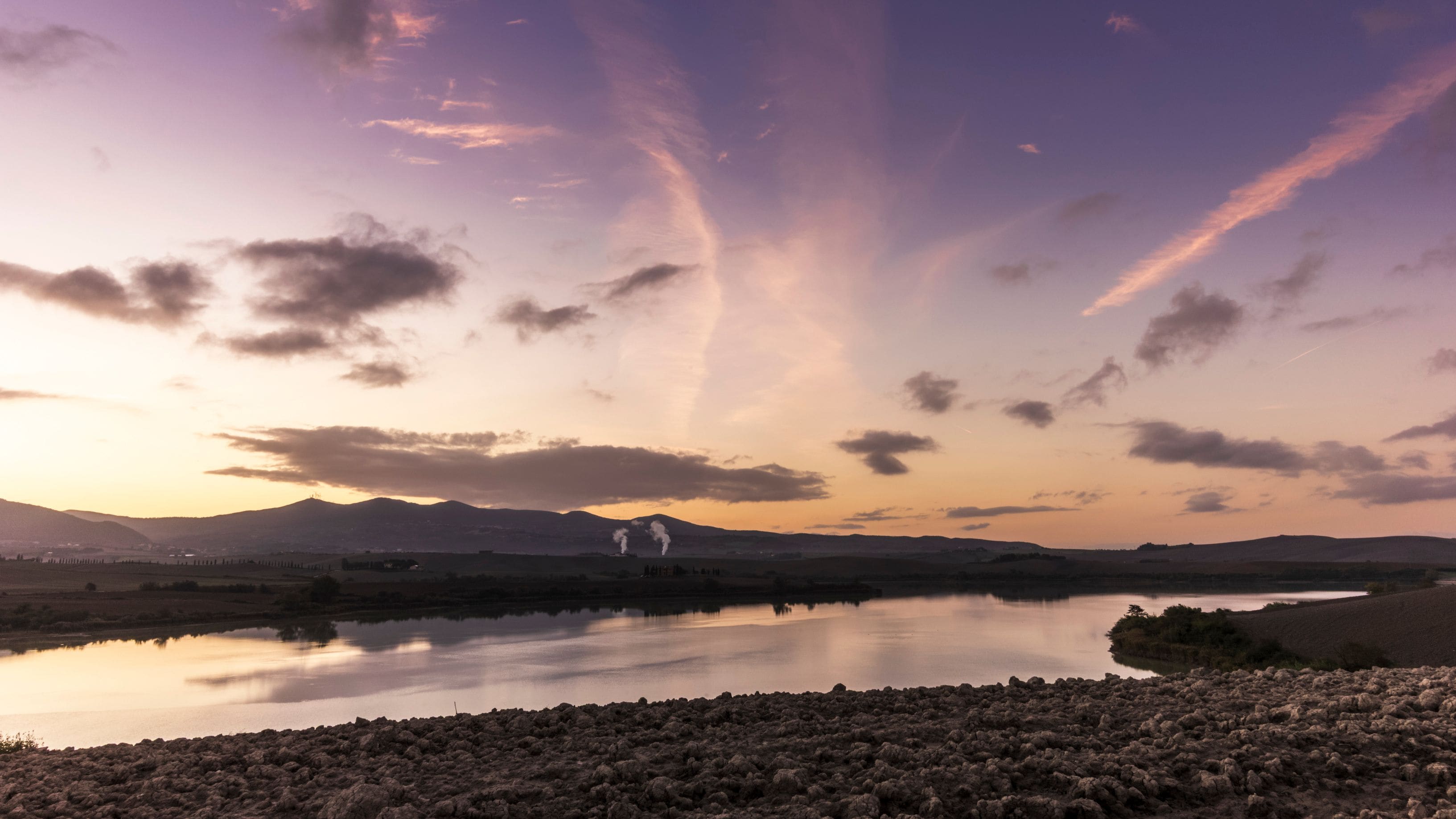 A sunset over a lake with mountains in the background and smoke rising from a structure.