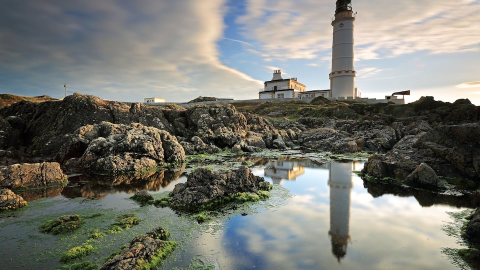 A lighthouse, rocky shoreline, and calm water reflecting the sky.