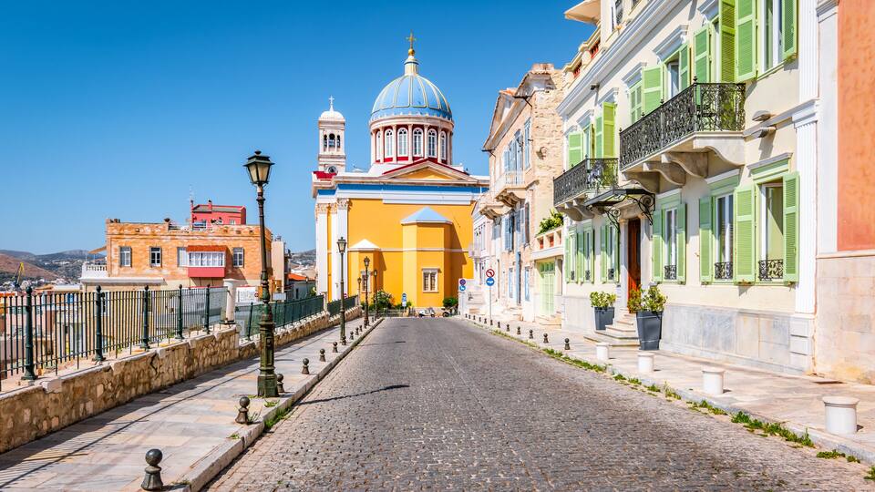 A cobblestone street lined with colorful buildings, a prominent yellow church with a blue dome, and a clear blue sky.