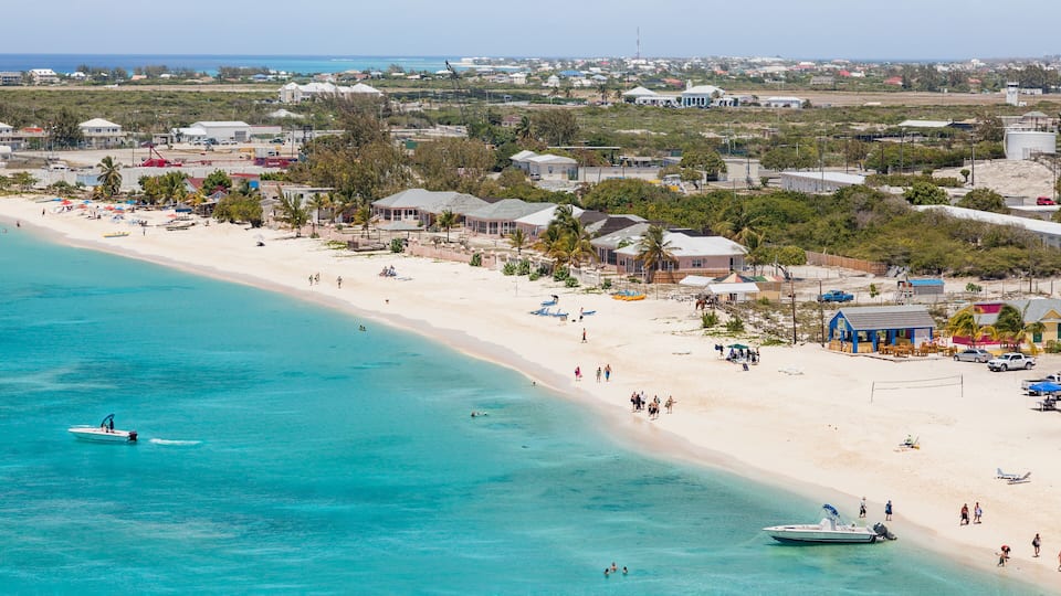 A beach with clear turquoise waters, a few boats, and a small town in the background.