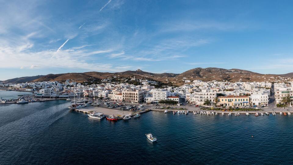 A coastal town with white buildings, a marina, and mountains in the background.
