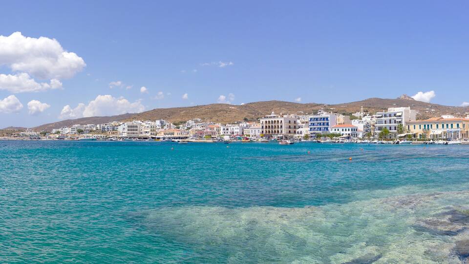 A coastal town with clear turquoise waters, white buildings, and a hill in the background.