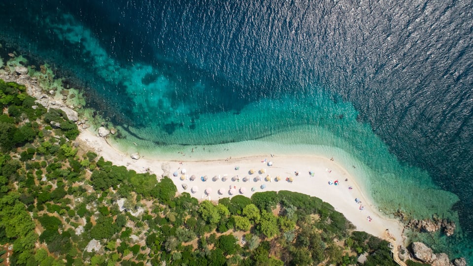 An aerial view of a beach with clear turquoise waters, a sandy shore, and lush greenery.