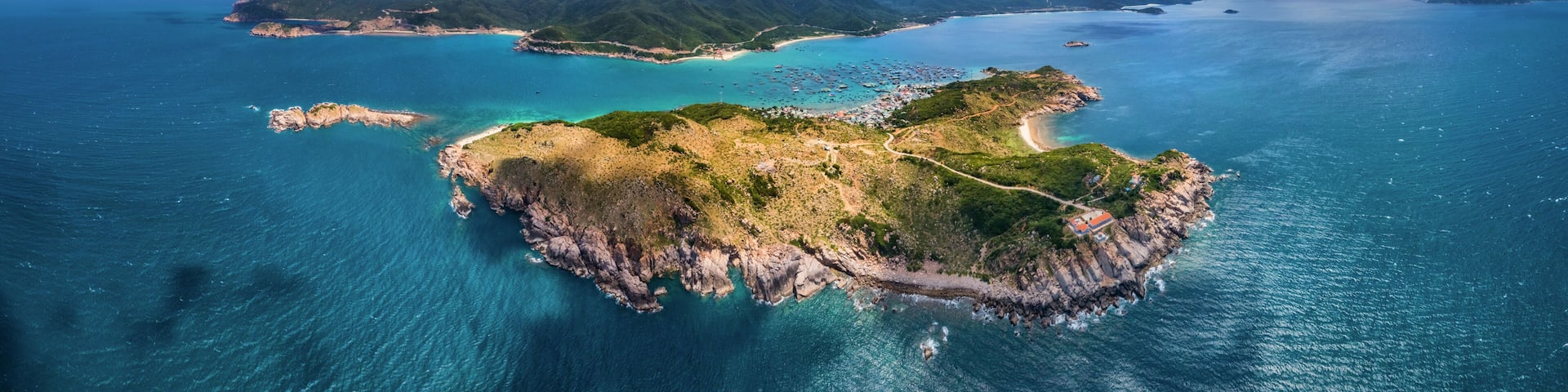 An aerial view of a coastal island with clear turquoise waters, green hills, and a small building on the shore.