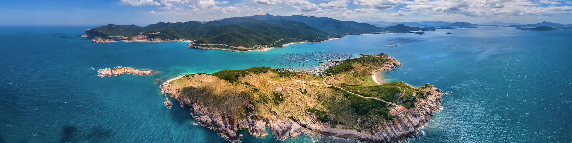 An aerial view of a coastal island with clear turquoise waters, green hills, and a small building on the shore.