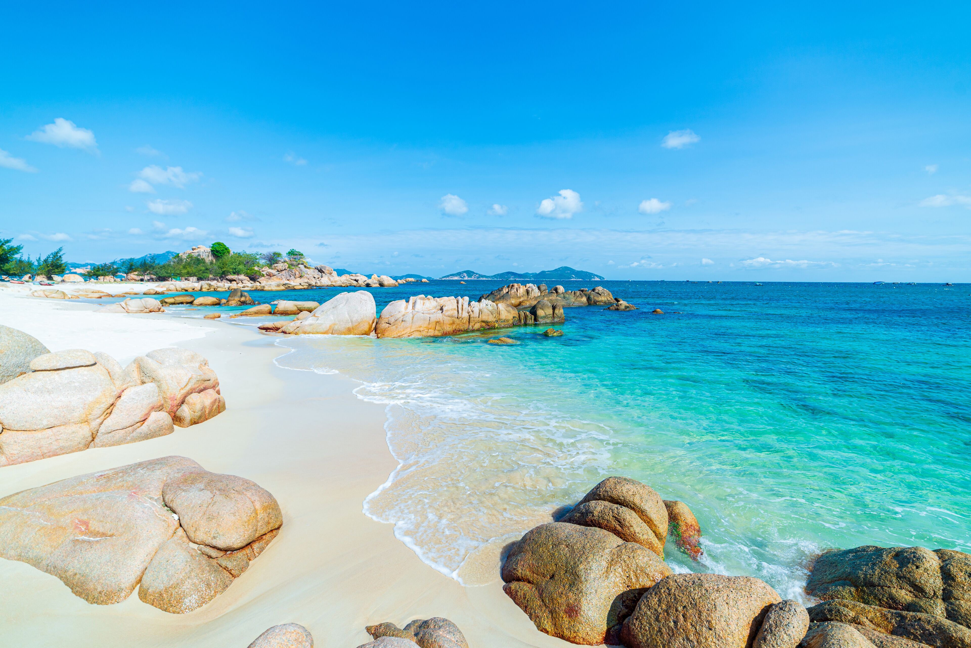 A beach with large rocks, clear turquoise water, and distant mountains.