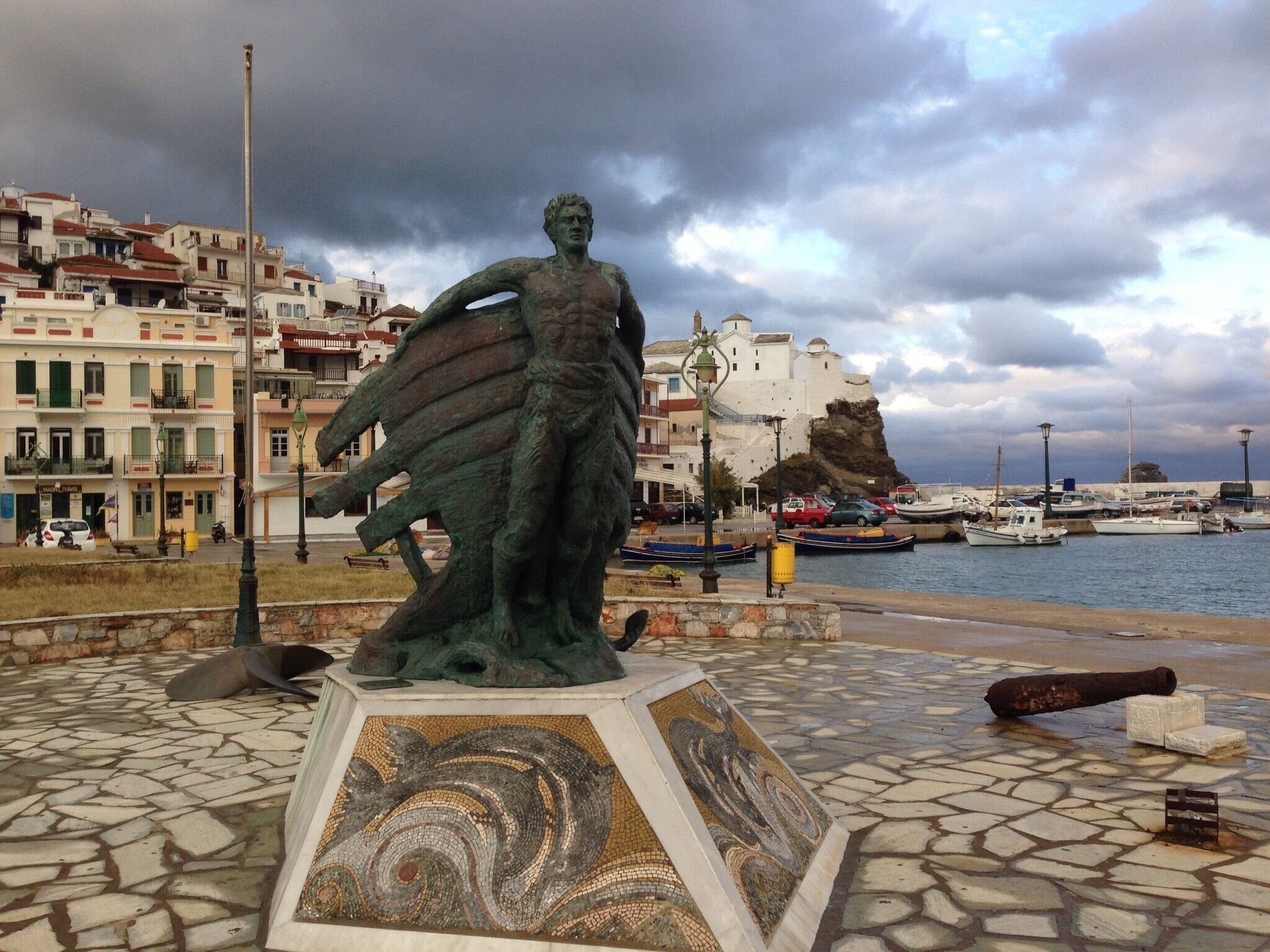 A statue of a figure with a sail, a mosaic base, and a rocky coastline with buildings in the background.