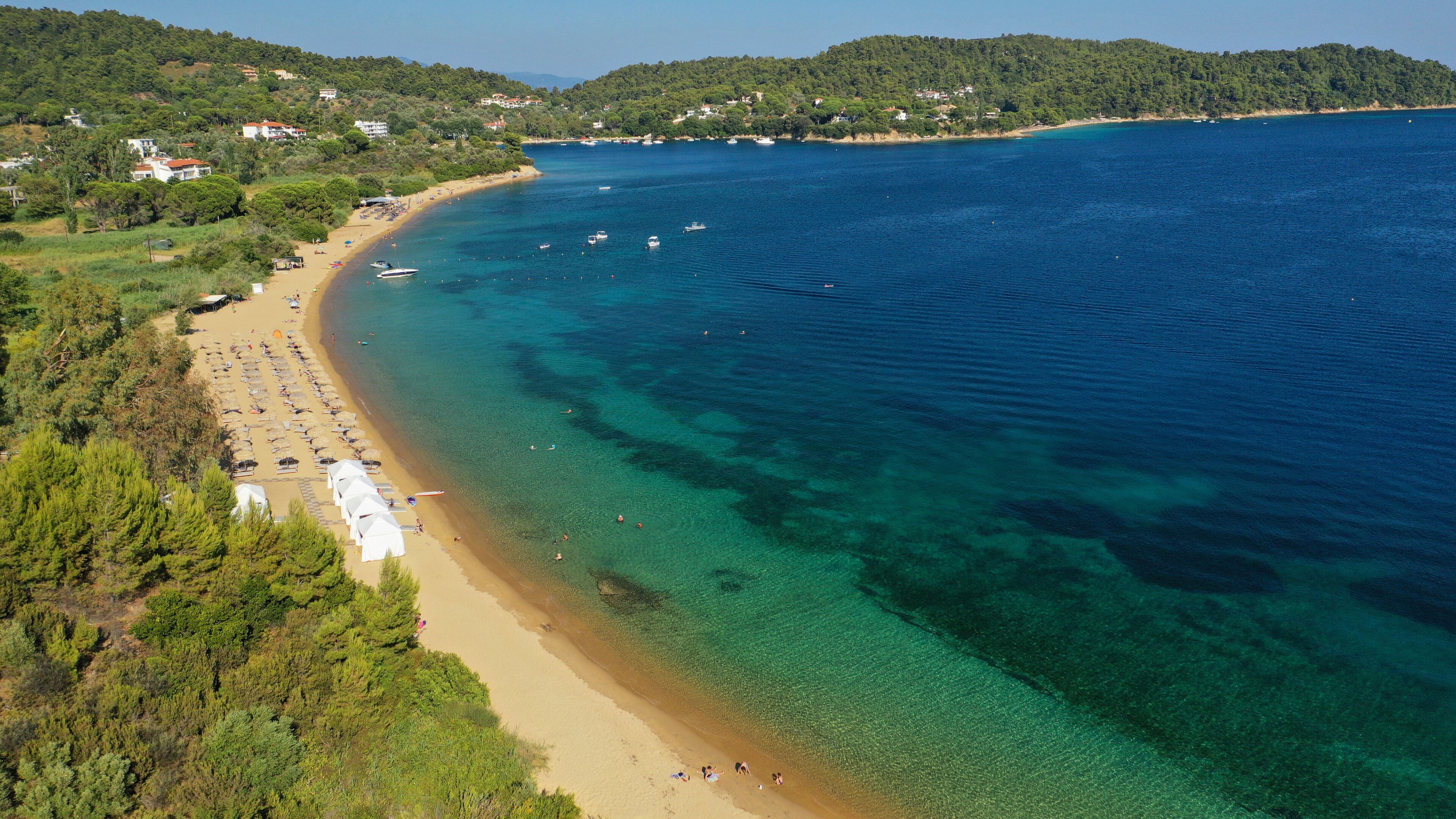 A coastal beach with clear waters, a sandy shore, and a backdrop of hills with scattered buildings.