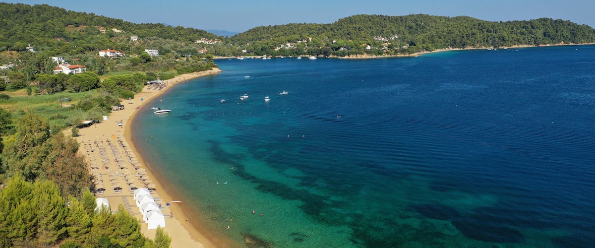 A coastal beach with clear waters, a sandy shore, and a backdrop of hills with scattered buildings.