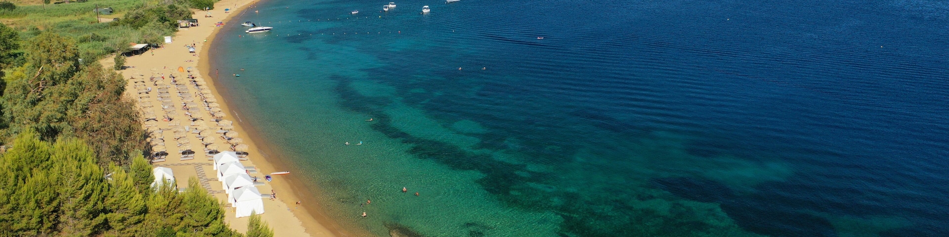 A coastal beach with clear waters, a sandy shore, and a backdrop of hills with scattered buildings.