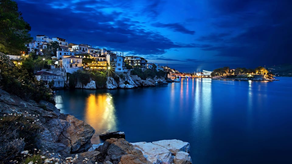 A coastal village at dusk with illuminated buildings, calm water, and rocky terrain.