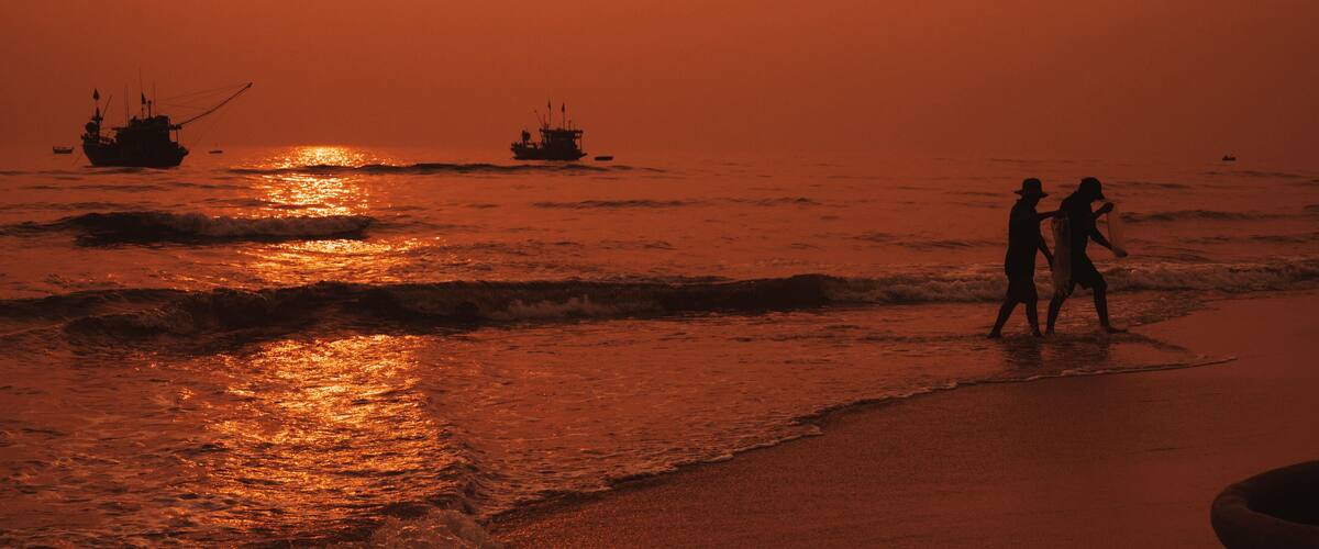 A sunset over the ocean with two people walking on the beach and several boats in the water.
