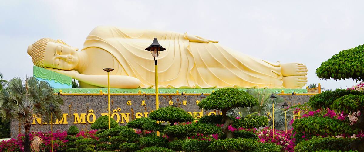 A large reclining Buddha statue with a golden robe, surrounded by lush greenery and vibrant flowers.