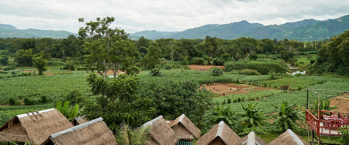 A rural landscape with thatched-roof huts, lush greenery, and distant mountains.