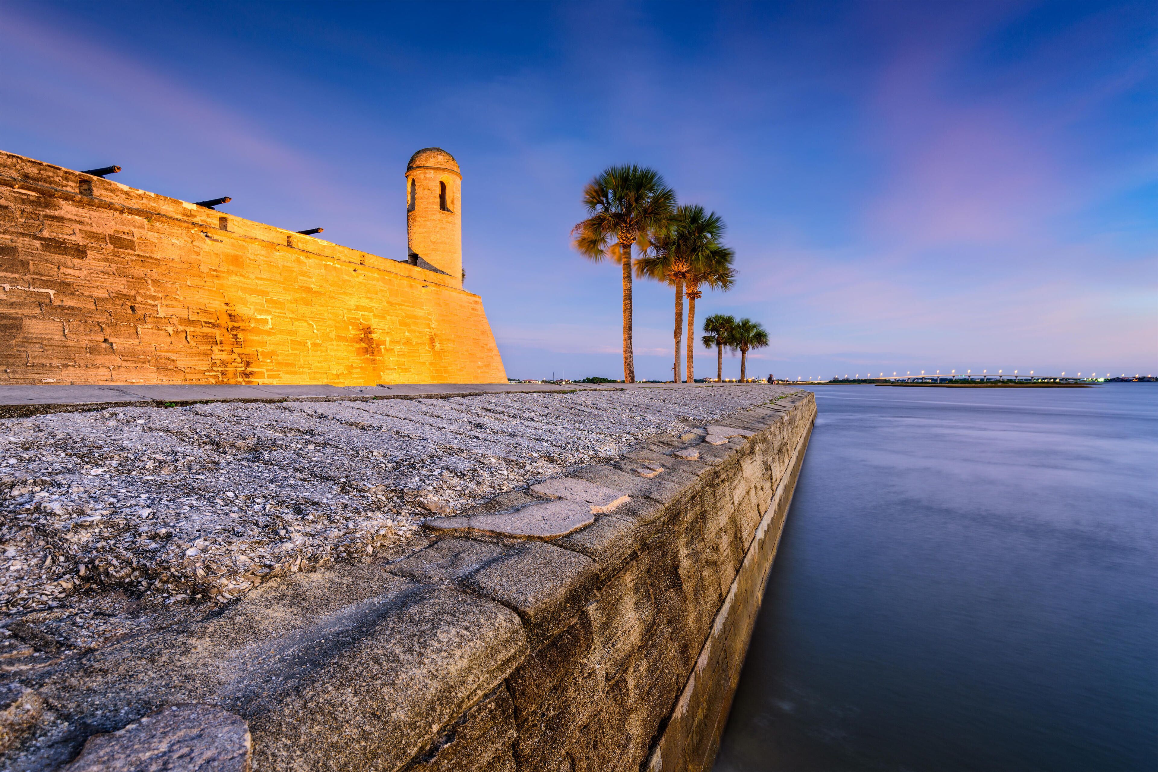 A stone wall with a tower, palm trees, and calm water.
