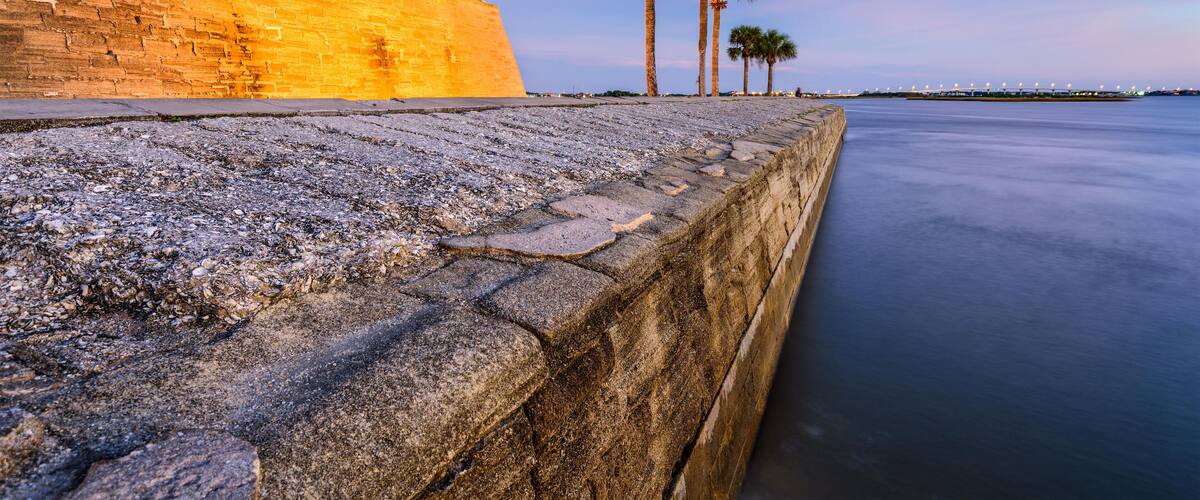 A stone wall with a tower, palm trees, and calm water.
