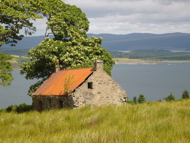 Una casa de piedra con techo oxidado, rodeada de hierba y un gran árbol, con vista a un lago y colinas lejanas.